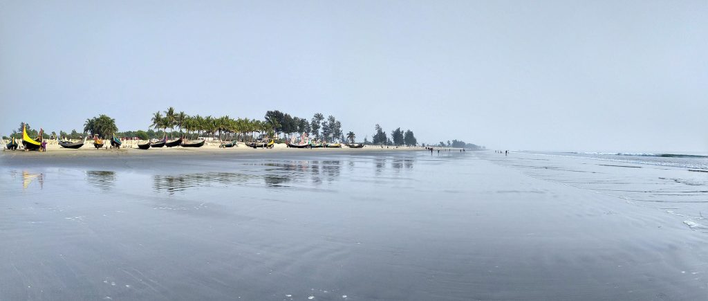 Scenic view of Cox's Bazar beach with fishing boats, trees, and the ocean under a gray sky.