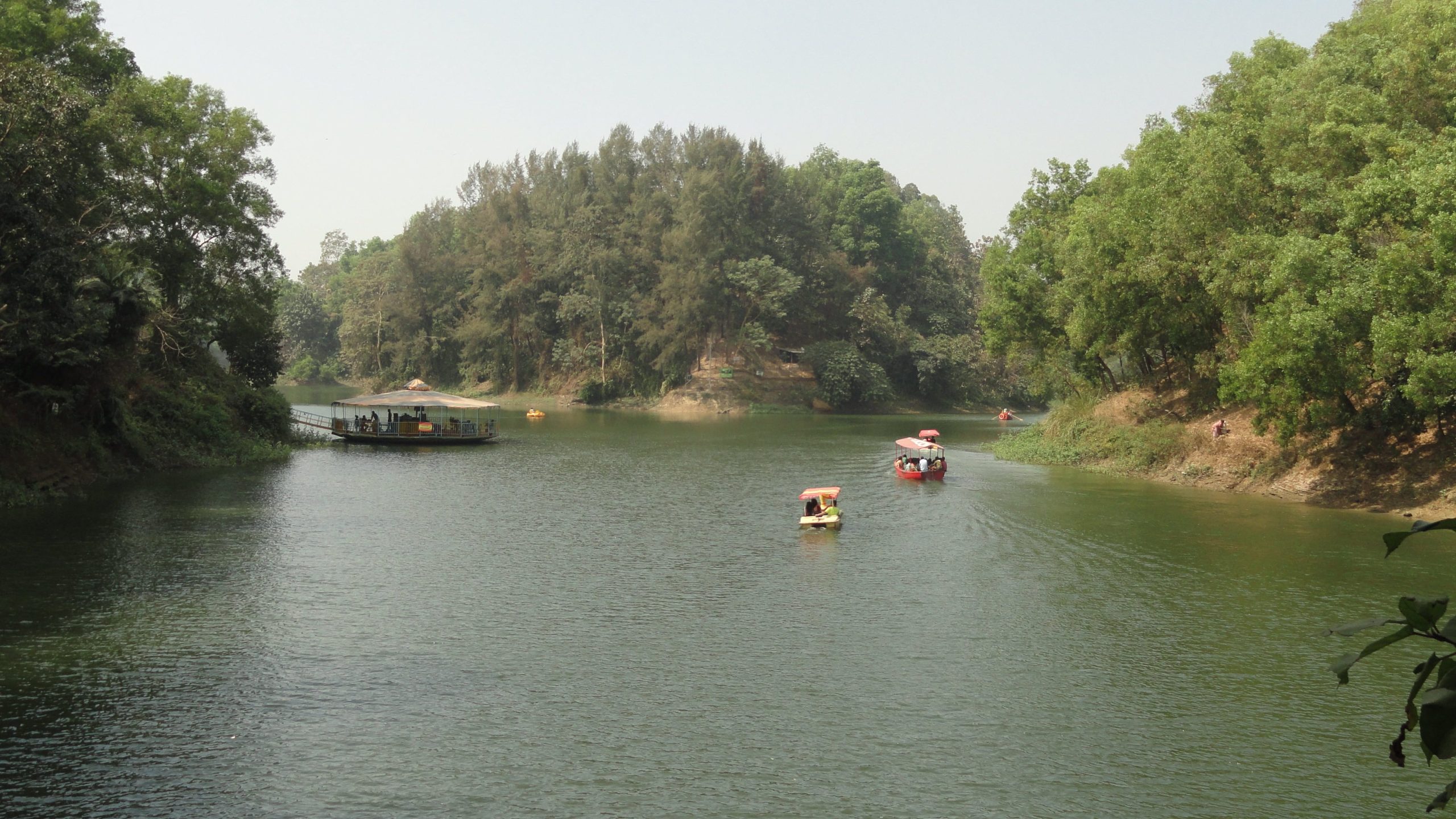 A calm lake scene at Foy's Lake, Chattogram, Bangladesh, featuring several small tour boats carrying passengers across the still water, with densely forested hills rising on either side under a light sky.