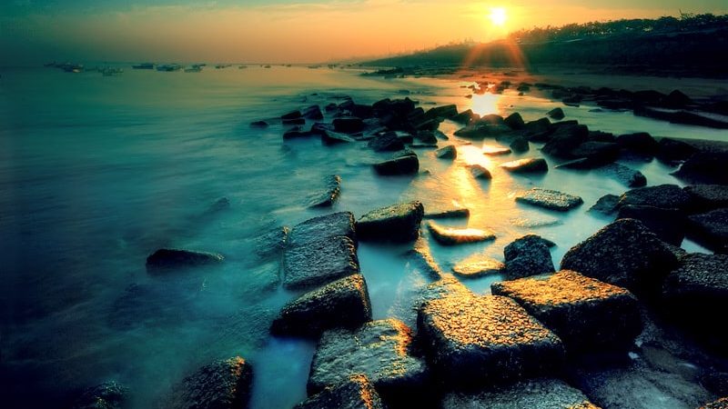 A vibrant sunset at Patenga Sea Beach, Chattogram, Bangladesh, featuring the unique arrangement of large, rectangular stones forming a breakwater, with the calm sea reflecting the golden light and several ships visible on the distant horizon.